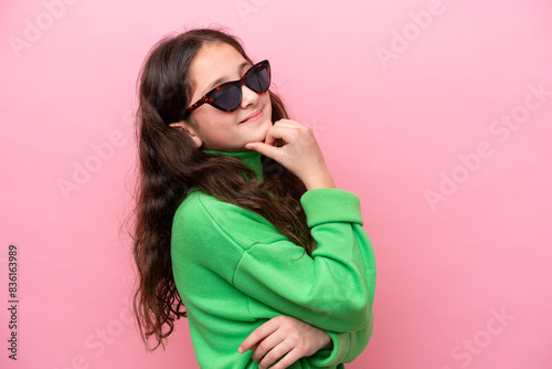 Little caucasian girl wearing sunglasses isolated on pink background looking side © luismolinero