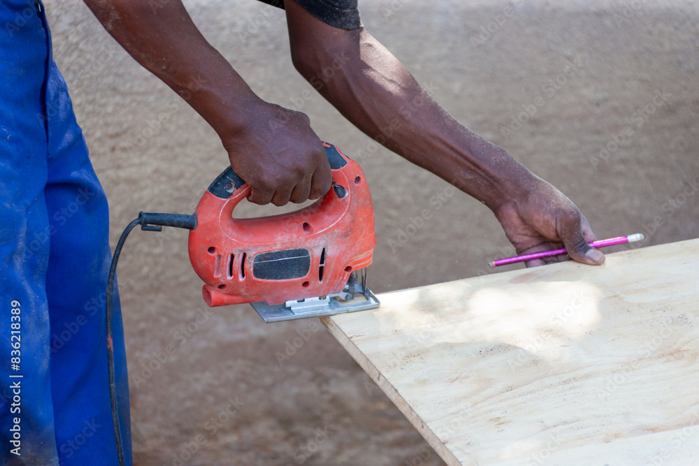 african man using a jigsaw to cut a plank of wood, holding a pencil to mark african man using a jigsaw to cut a plank of wood, holding a pencil to mark
