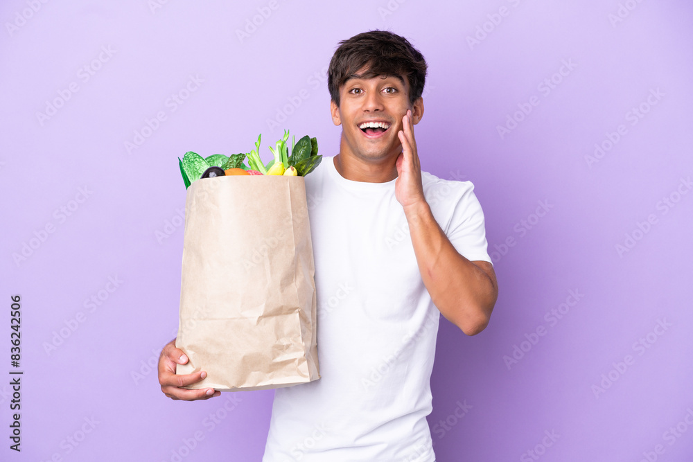 Young man holding a grocery shopping bag isolated on purple background with surprise and shocked facial expression Young man holding a grocery shopping bag isolated on purple background with surprise and shocked facial expression