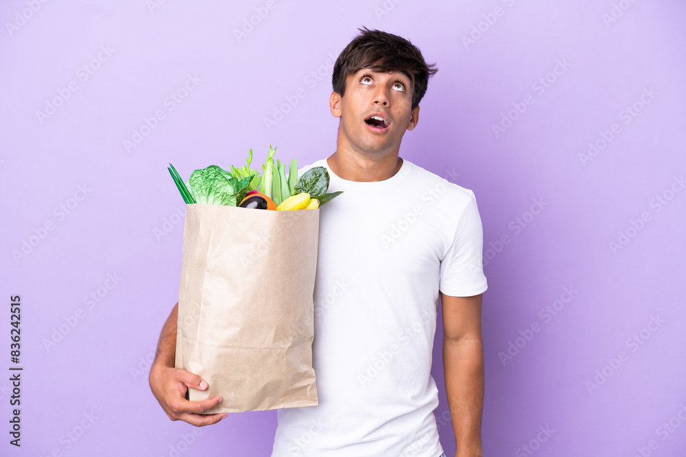 Young man holding a grocery shopping bag isolated on purple background looking up and with surprised expression Young man holding a grocery shopping bag isolated on purple background looking up and with surprised expression