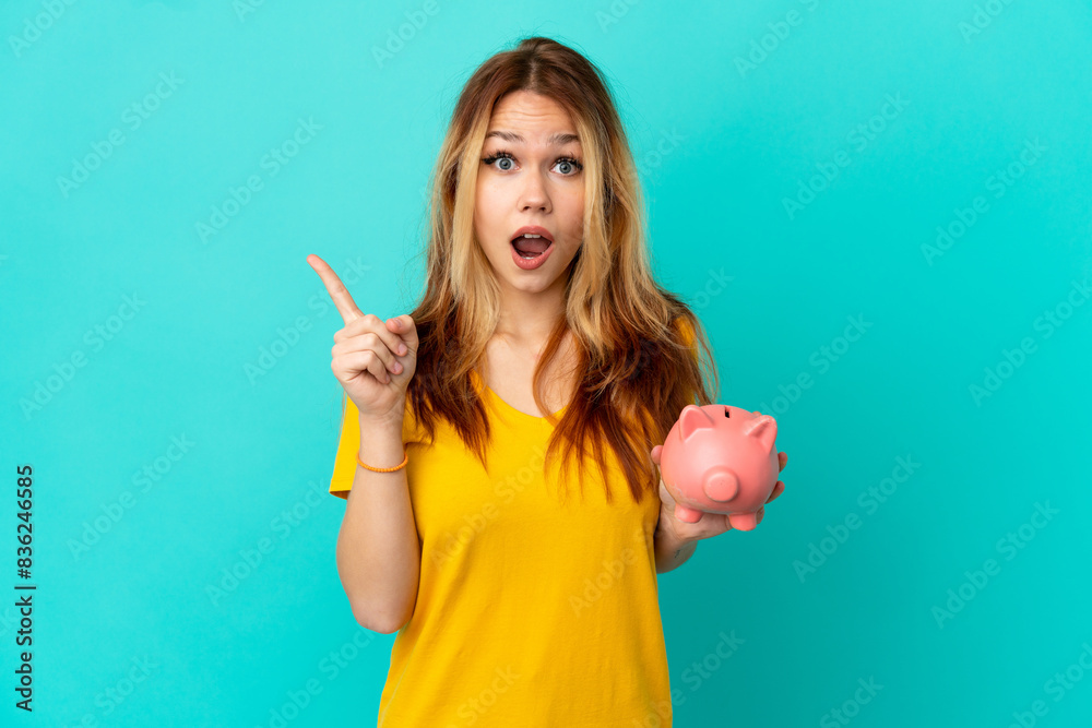 Teenager blonde girl holding a piggybank over isolated blue background surprised and pointing side Teenager blonde girl holding a piggybank over isolated blue background surprised and pointing side