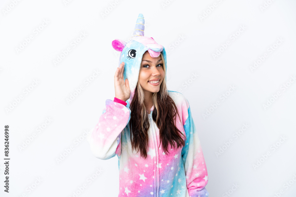 Young Russian woman wearing a unicorn pajama isolated on white background listening to something by putting hand on the ear Young Russian woman wearing a unicorn pajama isolated on white background listening to something by putting hand on the ear