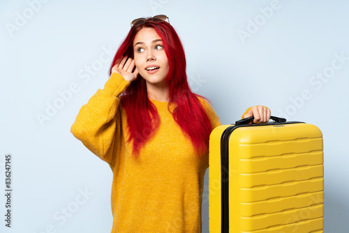 Traveler girl holding a suitcase isolated on blue background listening to something by putting hand on the ear