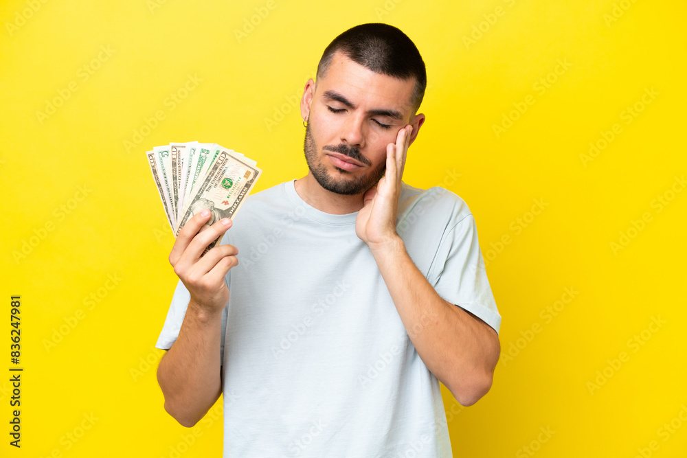 Young caucasian man taking a lot of money isolated on yellow background with headache Young caucasian man taking a lot of money isolated on yellow background with headache
