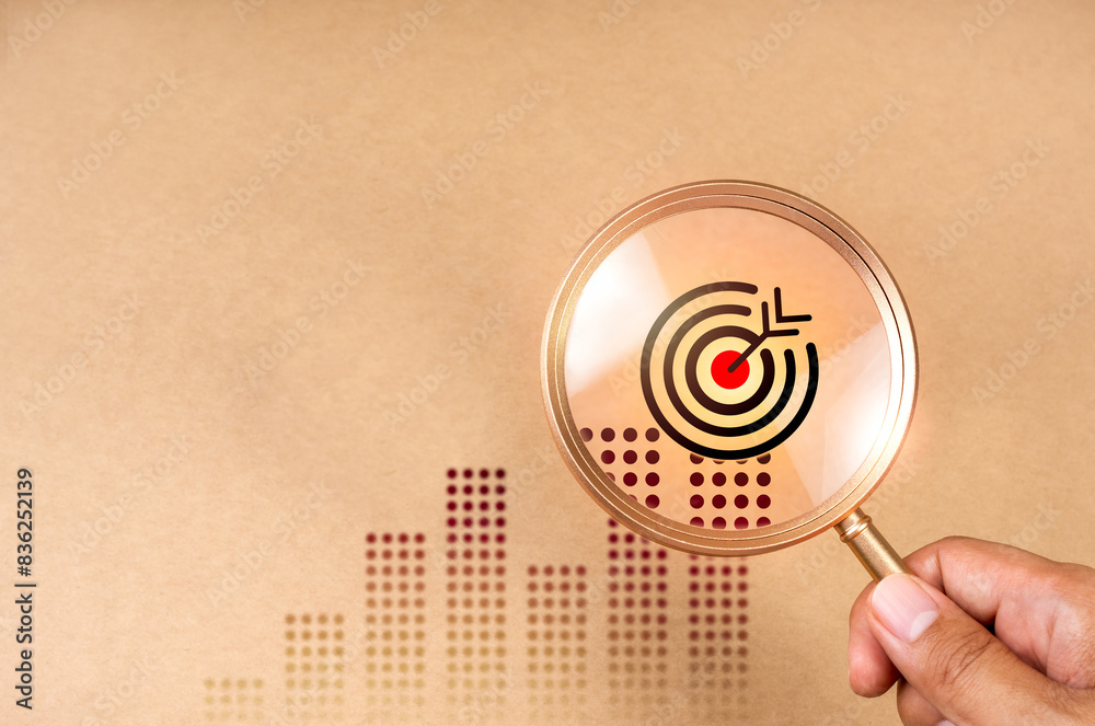 Business target growth concept. Bullseye dartboard icon on business graph expanded in magnifying glass lens in hand for focus business goal trends, brown background with copy space, minimal and eco. Business target growth concept. Bullseye dartboard icon on business graph expanded in magnifying glass lens in hand for focus business goal trends, brown background with copy space, minimal and eco.