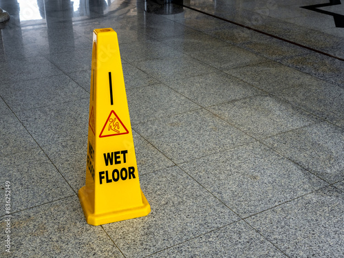 Wet floor caution sign on walkway in modern building. Warning red slip and fall people and text "WET FLOOR", sign on yellow plastic rubber cone on granite floor inside the building with copy space.