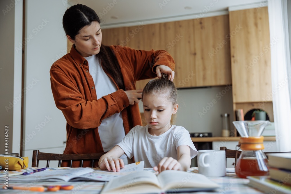 Mom combing her daughter's hair while she does her homework at home at the kitchen table Mom combing her daughter's hair while she does her homework at home at the kitchen table
