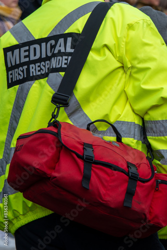 Medical health responder standing with their hands on their hips. Back of their yellow jacket has a medical first responder in grey reflective letters against black backing, a grey cross and armbands
