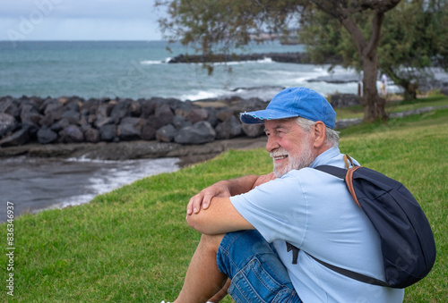 Smiling senior man sitting on the grass at the beach in a cloudy day enjoying free time vacation or retirement. Relaxed man wearing blue cap looking at the horizon over sea #836346937