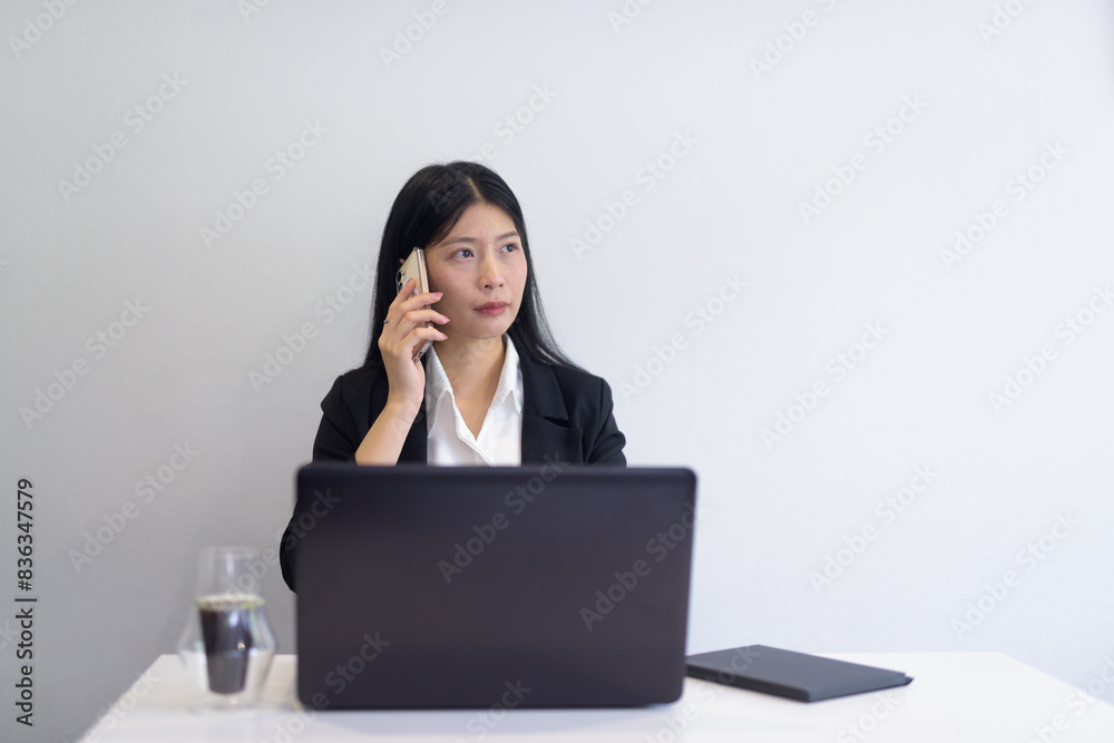 Asian businesswoman with laptop working in office, coffee and notebooks on desk. Professional setting while talking on phone.