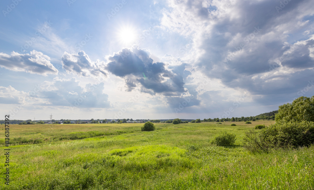 A sprawling green field stretches out under a bright blue sky, dotted with fluffy white clouds. The sun peeks through the clouds, casting a warm glow on the landscape.