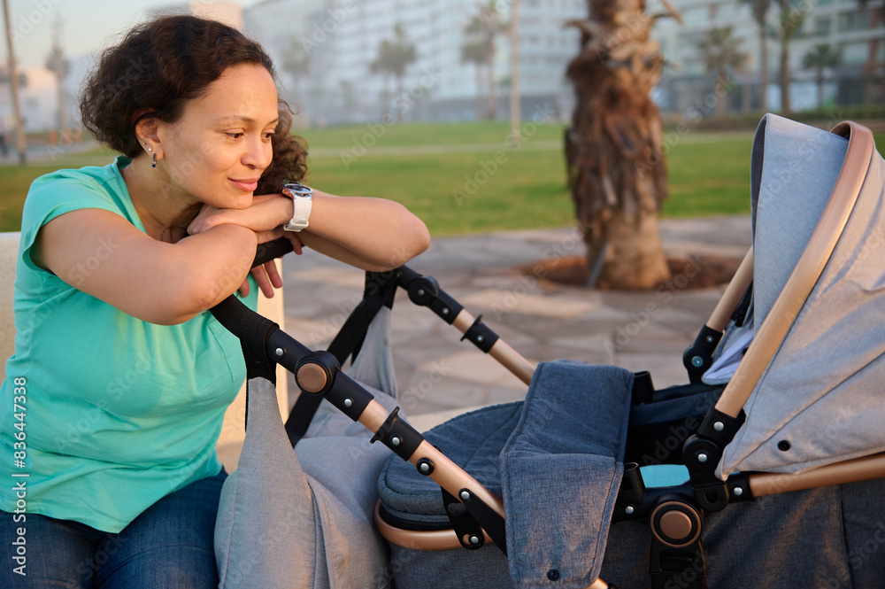 Obraz premium Beautiful multi ethnic young woman, mother 40s, smiling while admiring her baby sleeping in the baby pram, enjoying a happy weekend outdoors at sunset. People. Lifestyle. Leisure activity