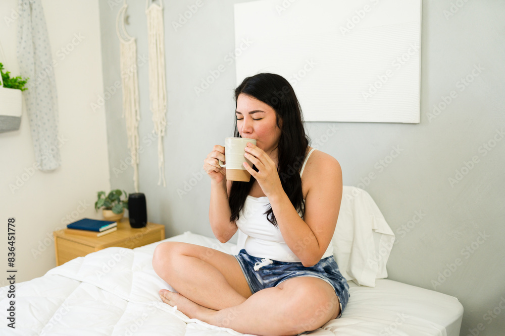 Cute woman enjoys a hot cup of coffee while sitting cross-legged on her bed in a serene bedroom scene Cute woman enjoys a hot cup of coffee while sitting cross-legged on her bed in a serene bedroom scene