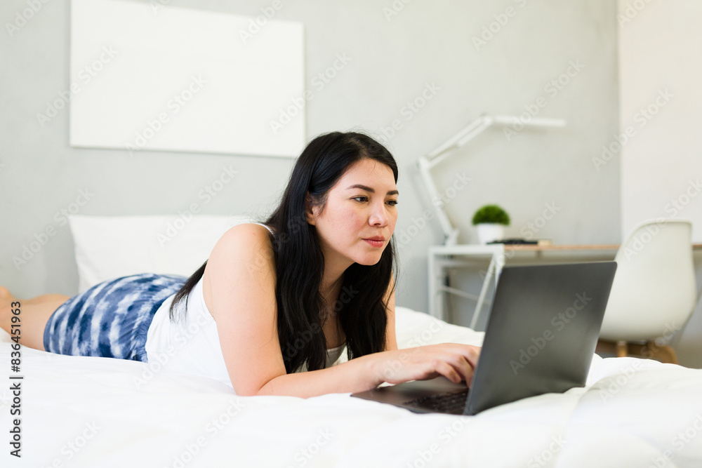 Focused woman lying on a bed while using a laptop in a bright and cozy modern bedroom setting Focused woman lying on a bed while using a laptop in a bright and cozy modern bedroom setting