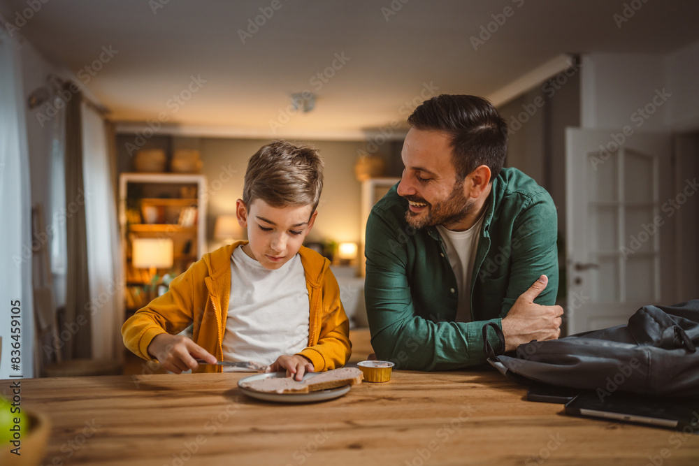Fototapeta premium Young boy smear pate on bread while dad pack his backpack for school