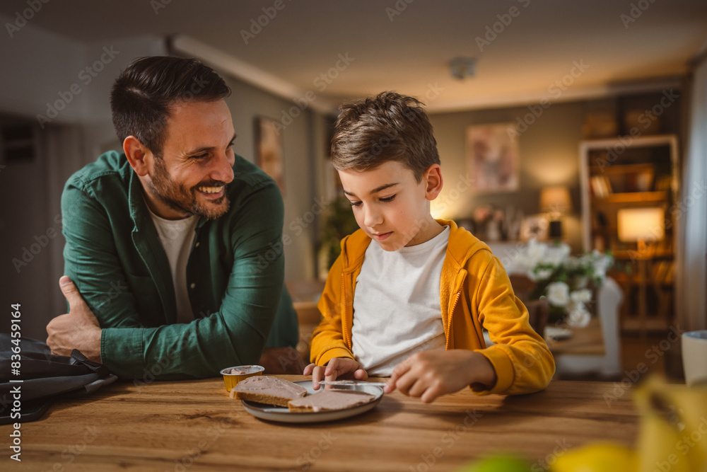 Young boy smear pate on bread while dad pack his backpack for school
