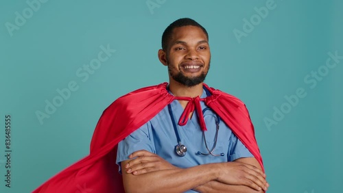 Smiling male nurse portraying superhero wearing red cape, isolated over studio background. Portrait of cheerful heroic medic posing as hero in costume with mantle, camera A
