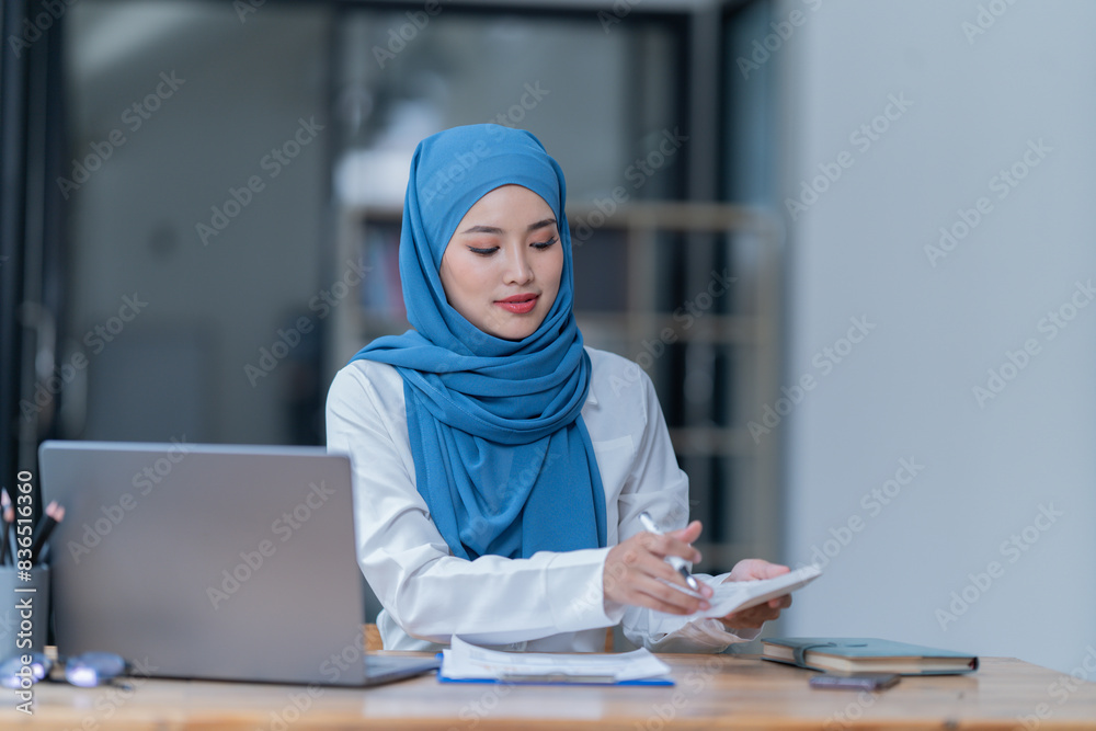 A female Arab entrepreneur is diligently working in her office, utilizing her laptop and documents to accomplish her tasks with contentment. A female Arab entrepreneur is diligently working in her office, utilizing her laptop and documents to accomplish her tasks with contentment.