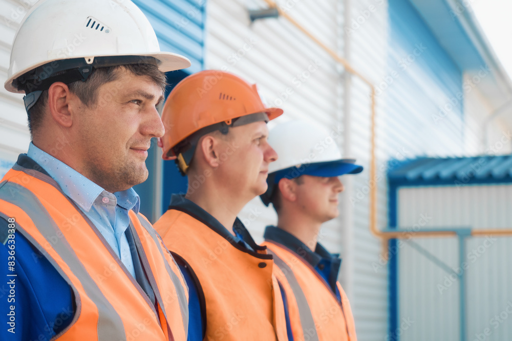 Three builders in helmets and vests stand in row. Friendly construction team of Caucasian appearance. Portraits of young workers at construction site on summer day. Three builders in helmets and vests stand in row. Friendly construction team of Caucasian appearance. Portraits of young workers at construction site on summer day.