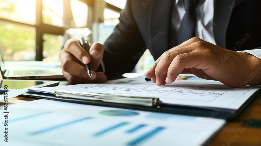 Businessman analyzing documents at a desk with a pen, laptop, and financial charts in a sunlit office. Businessman analyzing documents at a desk with a pen, laptop, and financial charts in a sunlit office.