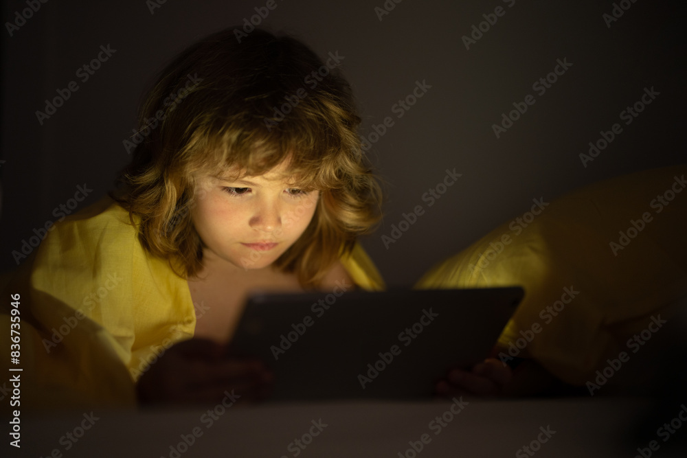 Bed time story. Child in bed under a blanket plays on a tablet in a game in the dark. The childs face is illuminated by a tablet. Child using tablet in bed at home. Kid in bedroom watching movie. Bed time story. Child in bed under a blanket plays on a tablet in a game in the dark. The childs face is illuminated by a tablet. Child using tablet in bed at home. Kid in bedroom watching movie.