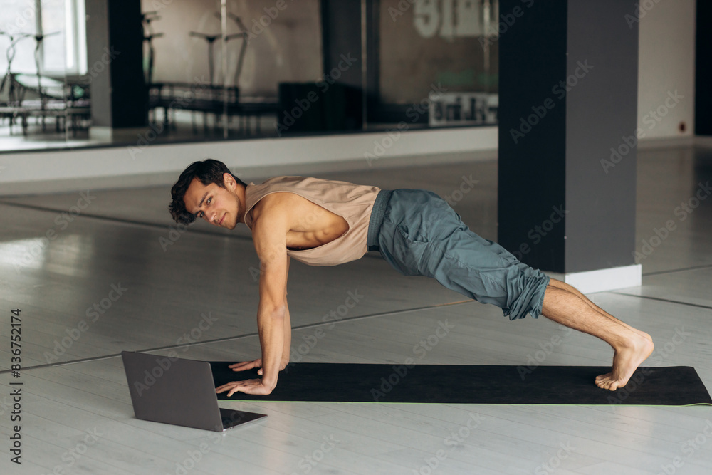 Fototapeta premium Young man exercising at home, lying in plank position and using laptop
