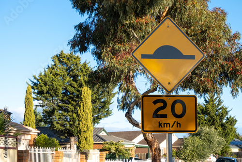A traffic warning sign indicates the presence of a speed hump on the road with a 20km/h speed restriction applied in Australia, with a blurry view of suburban houses in the neighborhood. photo