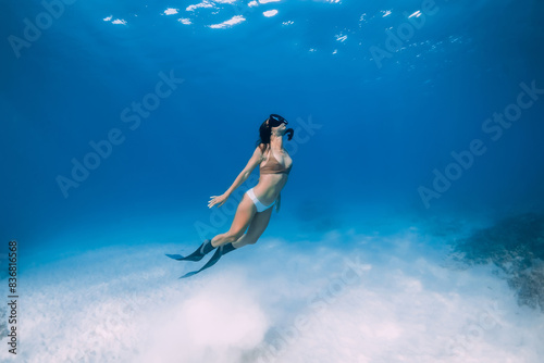 Female free diver swims underwater over sandy sea in blue ocean.