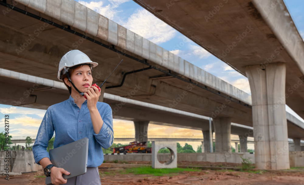 Project manager, female engineer wearing a helmet, holding a computer, using a walkie-talkie to work on the expressway bridge construction site. Project manager, female engineer wearing a helmet, holding a computer, using a walkie-talkie to work on the expressway bridge construction site.