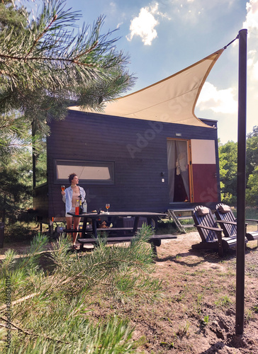 woman with a glass of cocktail resting near A modular frame-type apartment on wheels, lined with wood against the background of an summer forest.