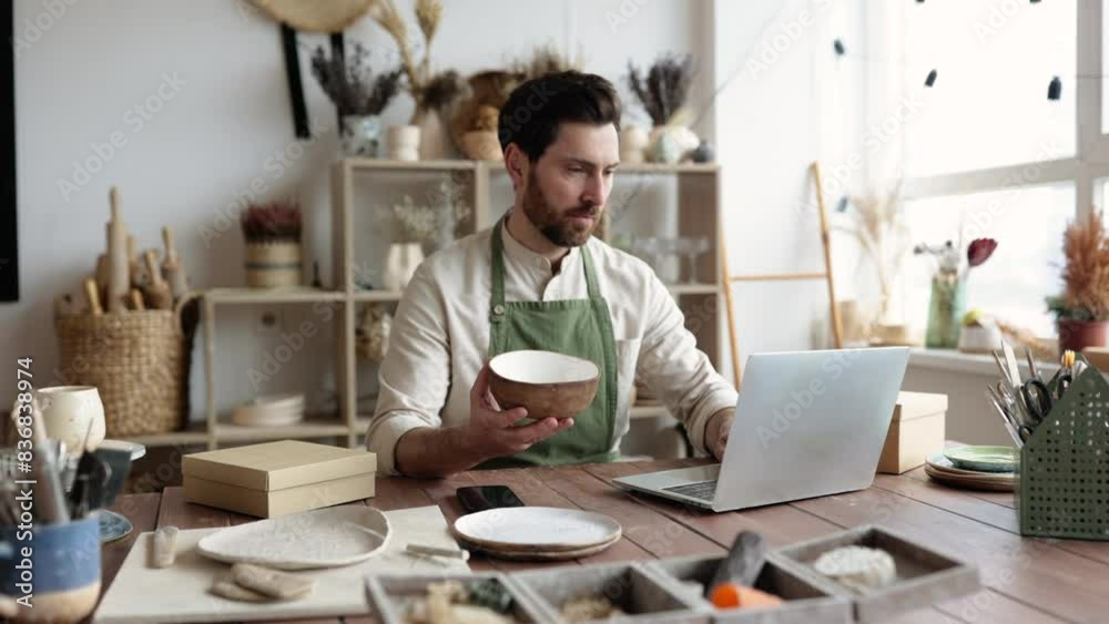 Adult ceramic shop owner using laptop while working in store. Male entrepreneur managing online order of mug on website. Confident young businessman running creative small business.