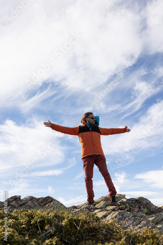 A man in an orange jacket stands on a rocky hillside, looking up at the sky