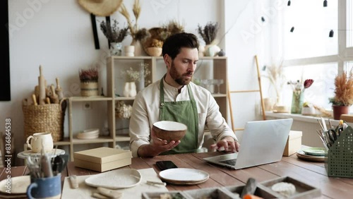 Adult worker in apron holds unique bowl created in personal style against background of shelves with dishes. Adult bearded male potter engaged in e-commerce selling handmade clay products on Internet. photo