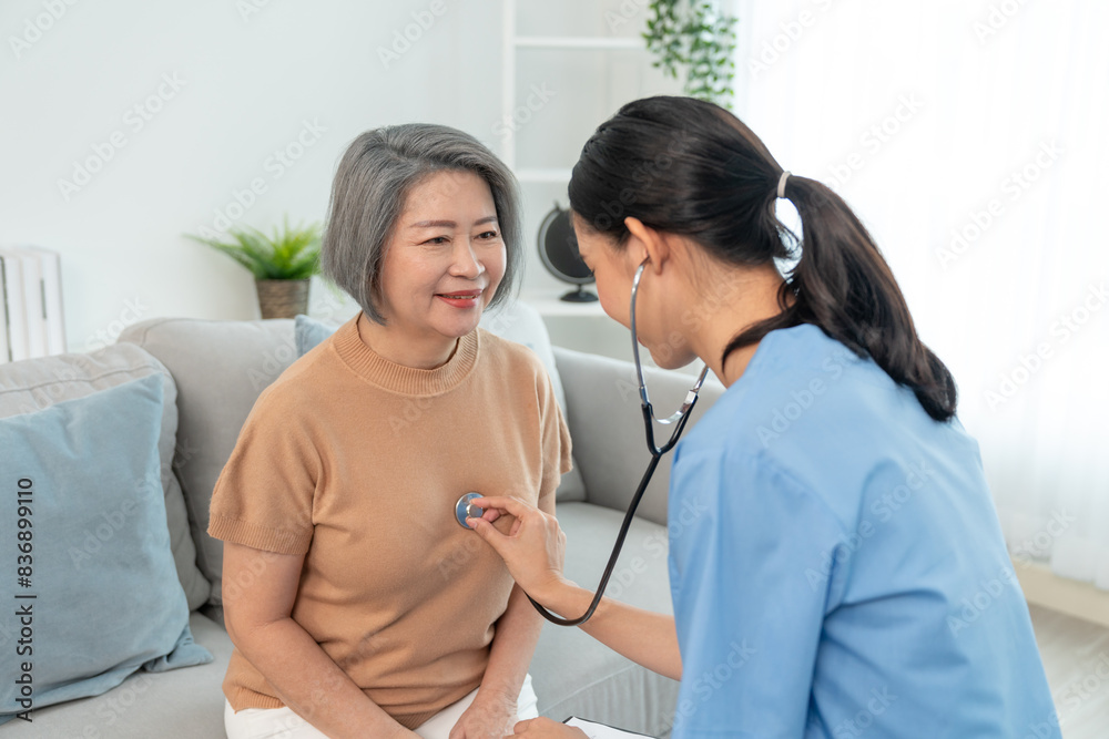 Asian senior elderly woman patient measures heartbeat by stethoscope while sitting on sofa in living room at home.Asian nurse woman caregiver during home visit. Asian senior elderly woman patient measures heartbeat by stethoscope while sitting on sofa in living room at home.Asian nurse woman caregiver during home visit.