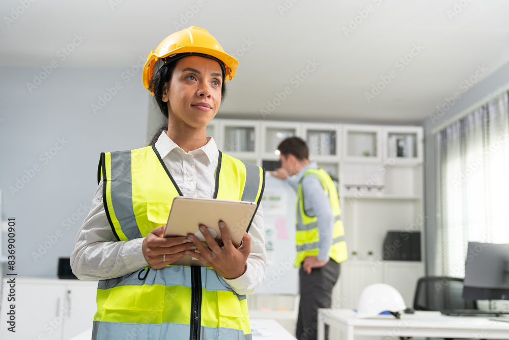 Portrait of confident female engineer standing and holding tablet at construction office. Woman foreman wearing vest and hardhat safety ready to team