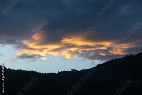 Landscape of thick clouds, orange light hitting the blue sky, silhouettes of trees, forests, and mountains. Area for text in the middle of the image