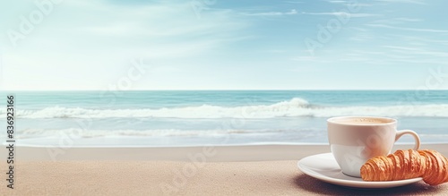 Woman holding a croissant and coffee cup on the beach with a copy space image included.