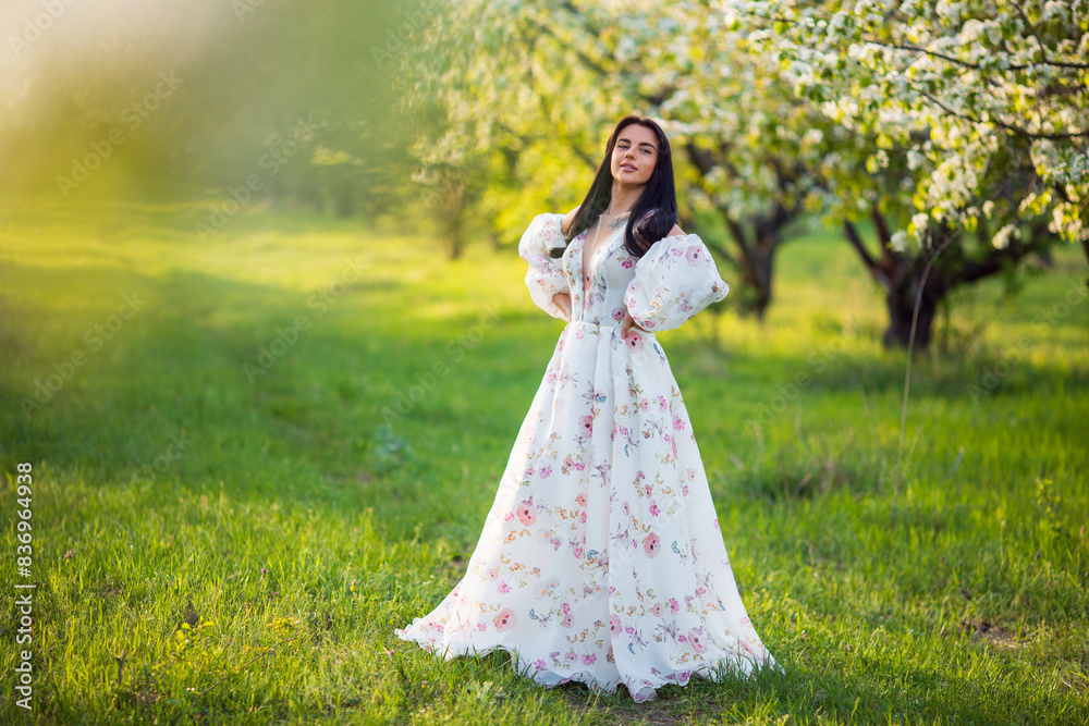 Full-length portrait of a beautiful brunette caucasian woman standing in blossoming apple garden in white floral dress on a fresh green grass and looking in the camera Full-length portrait of a beautiful brunette caucasian woman standing in blossoming apple garden in white floral dress on a fresh green grass and looking in the camera