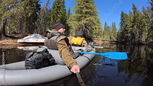 Two men are paddling down a calm river surrounded by trees. The weather is sunny and the water is clear. They are enjoying a relaxing day on the water, surrounded by nature.