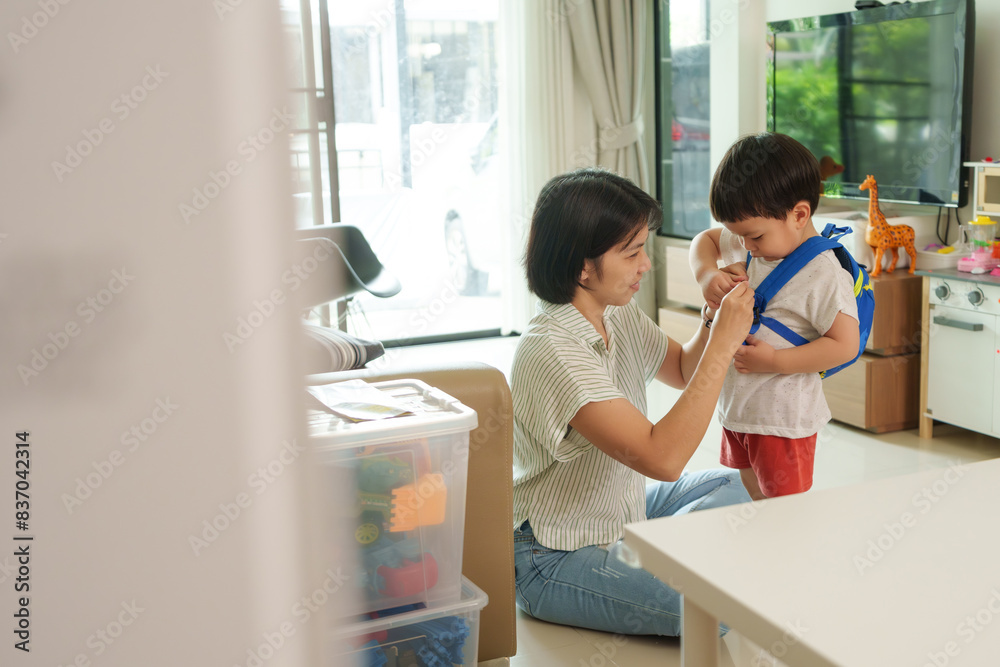 Asian Mother helps young son with blue backpack in living room. Child stands while mother adjusts the backpack. Morning routine, parent-child bonding, preparation for school. Asian Mother helps young son with blue backpack in living room. Child stands while mother adjusts the backpack. Morning routine, parent-child bonding, preparation for school.