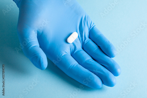 A single white pill placed in a blue-gloved hand against a soft blue background.