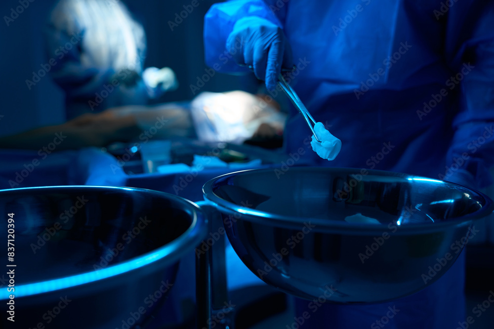 Doctor's hand in blue glove holding tweezers with cotton pad during operation Doctor's hand in blue glove holding tweezers with cotton pad during operation