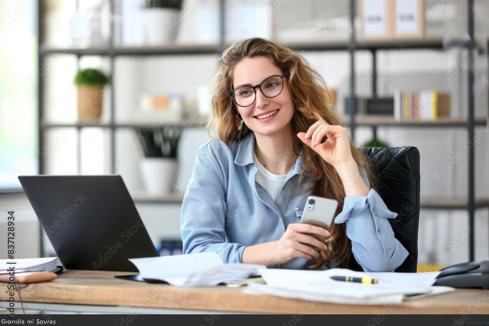 A woman is sitting at a desk with a laptop and a cell phone. She is smiling and holding the cell phone in her hand A woman is sitting at a desk with a laptop and a cell phone. She is smiling and holding the cell phone in her hand