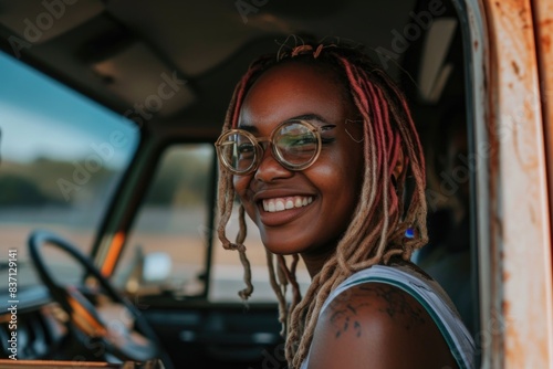 A woman with dreadlocks and glasses is smiling in a car. She is wearing a white tank top and has a tattoo on her arm