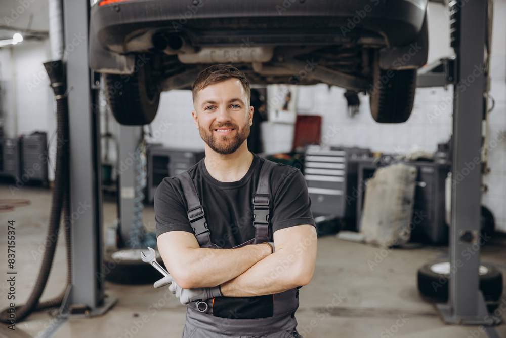 Smiling bearded mechanic while standing with crossed arms and holding wrenches in a car service