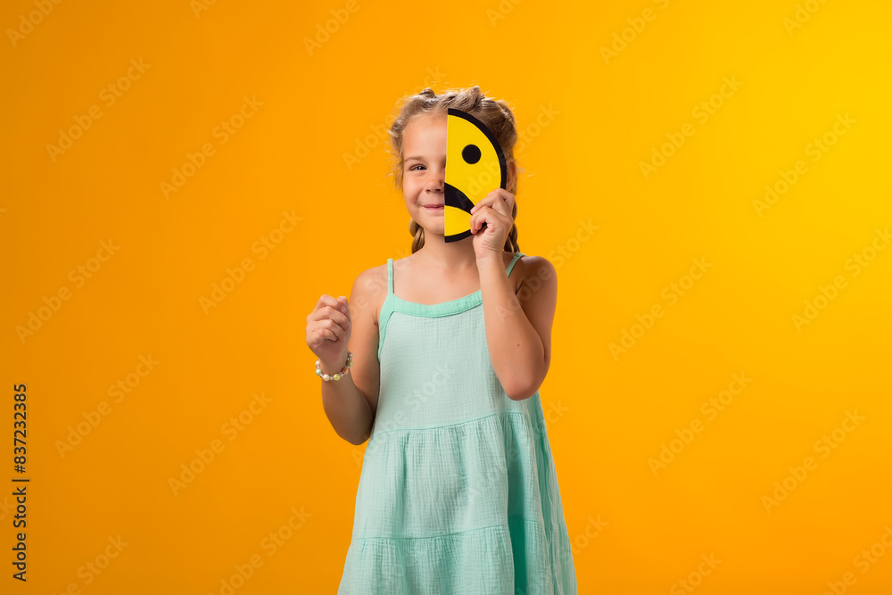 Fototapeta premium Smiling kid girl holding sad half face emoticon and showing thumbs up gesture. Mental health, psychology and children's emotions concept