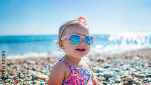 bebé femenino tierno y feliz en la arena de la playa con gafas de sol, mirando de frente a la camara 