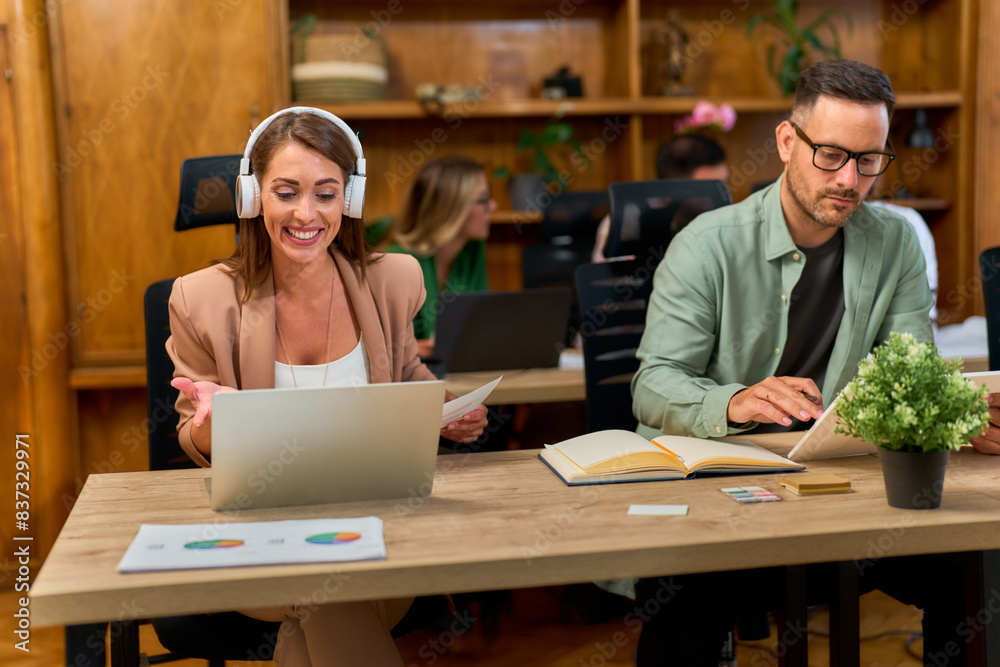 Fototapeta premium Two colleagues working in a modern office. A woman is smiling and wearing headphones while using a laptop, and a man is focused on his laptop and notebook.