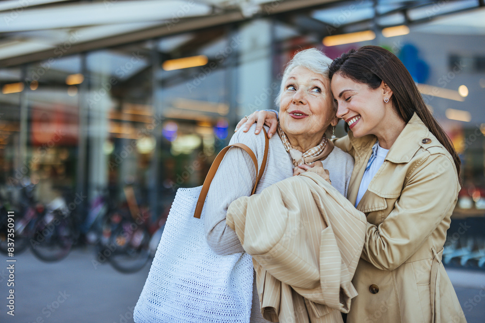 Fototapeta premium A young woman smiling supports her elderly mother, both dressed casually, as they carry grocery bags together outside a supermarket, highlighting family care and elder assistance.