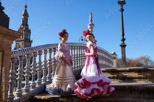 Two pretty little girls dancing flamenco dressed in typical gypsy costume pose on the stairs in their dress in a famous square in seville. The girls are happy. Flamenco, cultural heritage of humanity.
